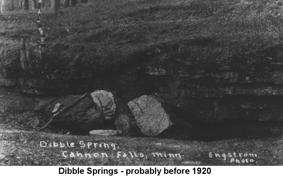 Black and white photo of large boulders in a crevice through which flows a stream; a high stone and earthen bank stands above the water. Handwritten in white ink on the photo is 'Dibble Spring, Cannon Falls, Minn. Engstrom Photo.